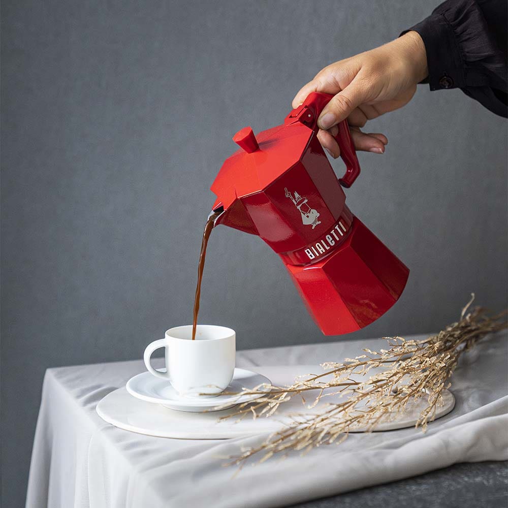 Hand pouring dark roast coffee from a red Bialetti moka pot into a white cup and saucer on a light tablecloth