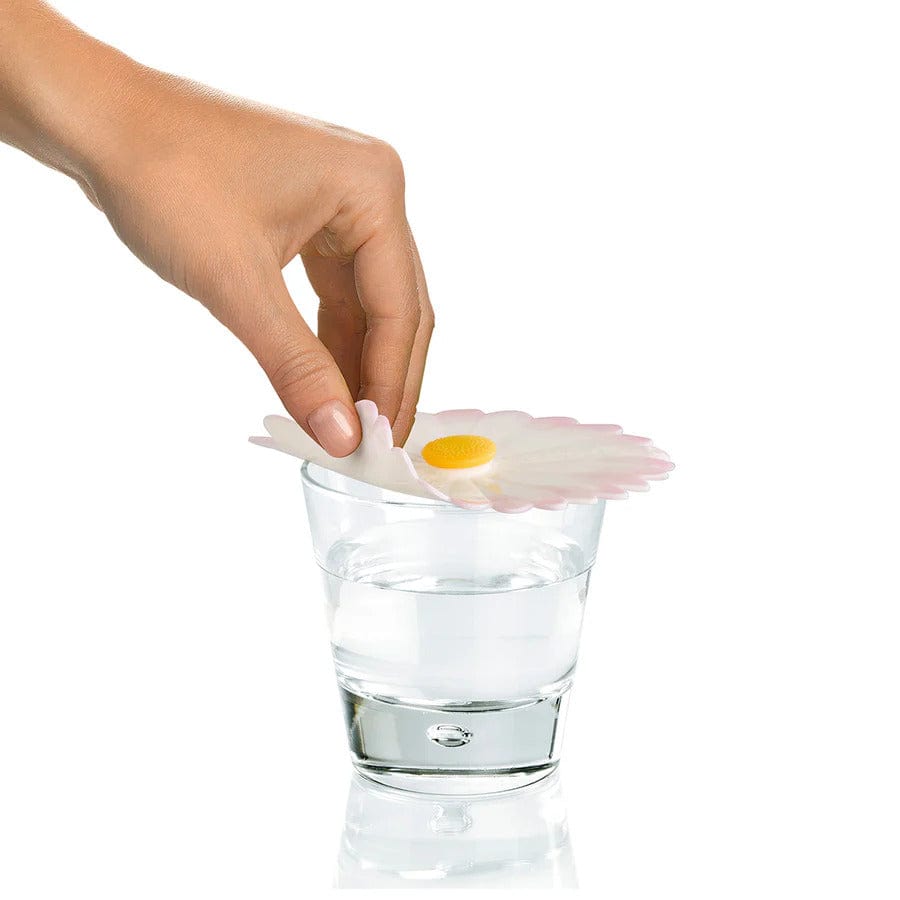 Hand placing a white and pink daisy-shaped silicone drink cover with a yellow center onto a glass of water