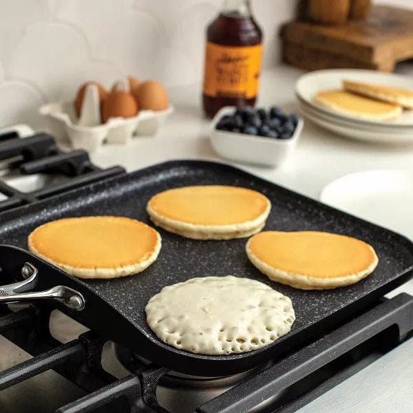 Black Nordic Ware Basalt 11 Square Griddle on a stovetop with pancakes cooking surrounded by breakfast items