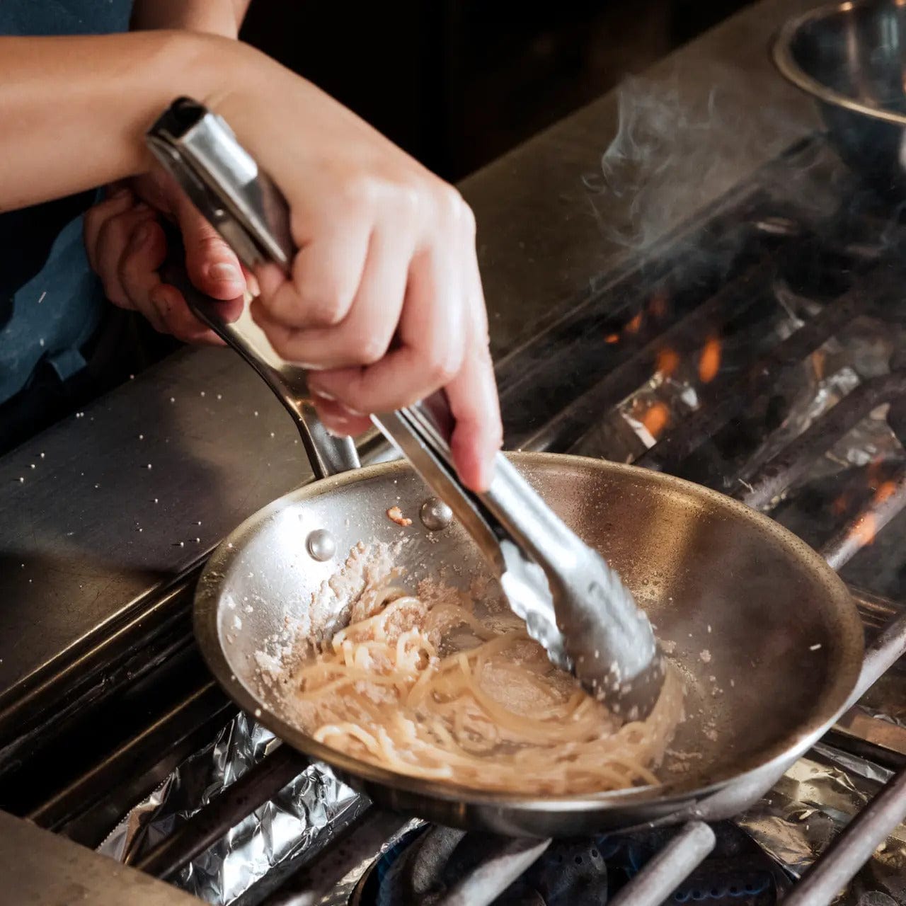 A chef stirring pasta in a shiny 10 stainless clad frying pan on a stovetop burner