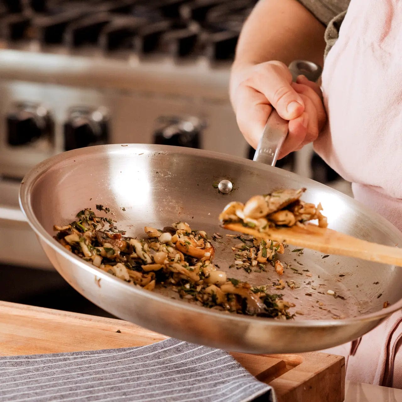 A hand holds a 12-inch stainless steel clad frying pan with sauteed vegetables