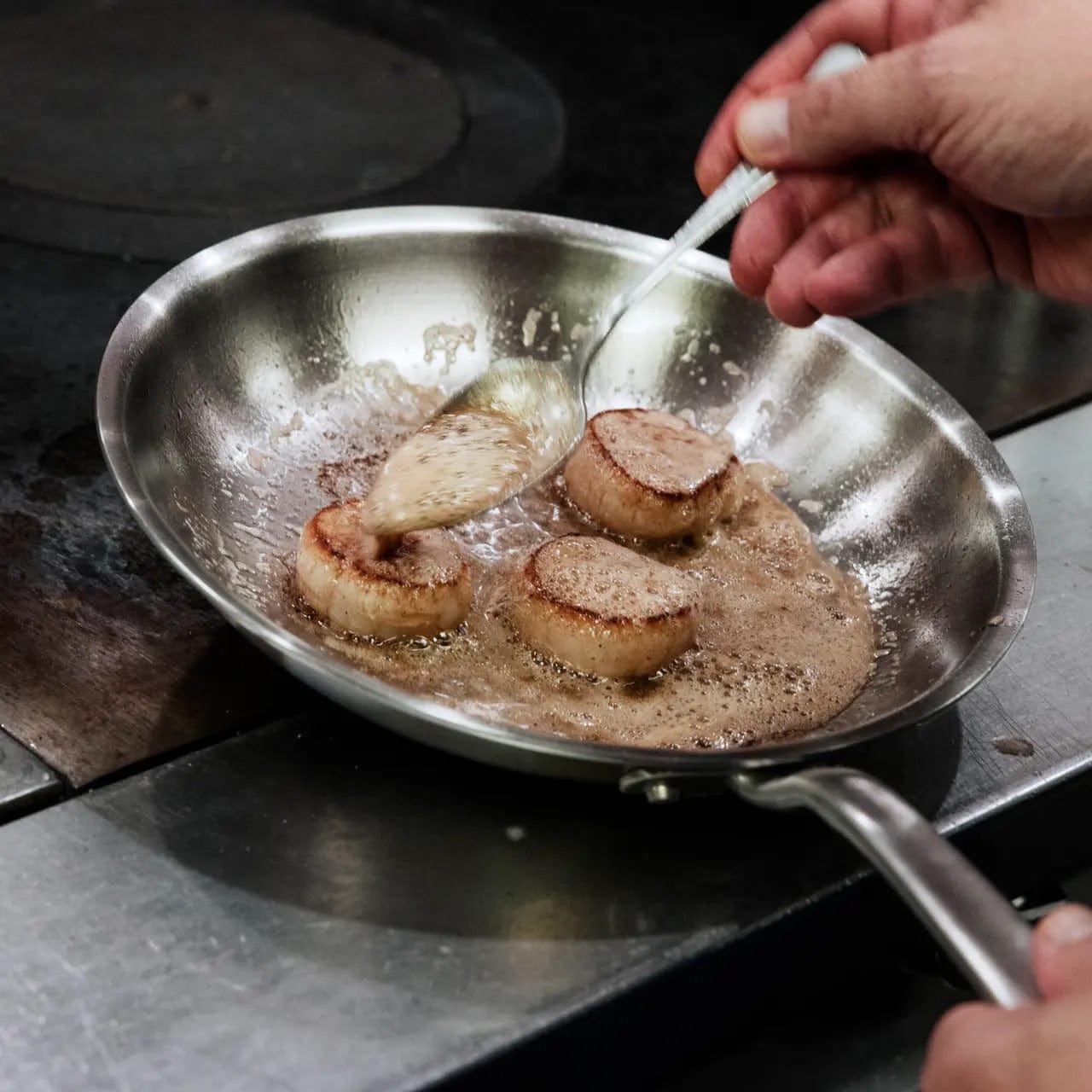 8 stainless clad frying pan on a stovetop with scallops searing in butter A hand bastes them