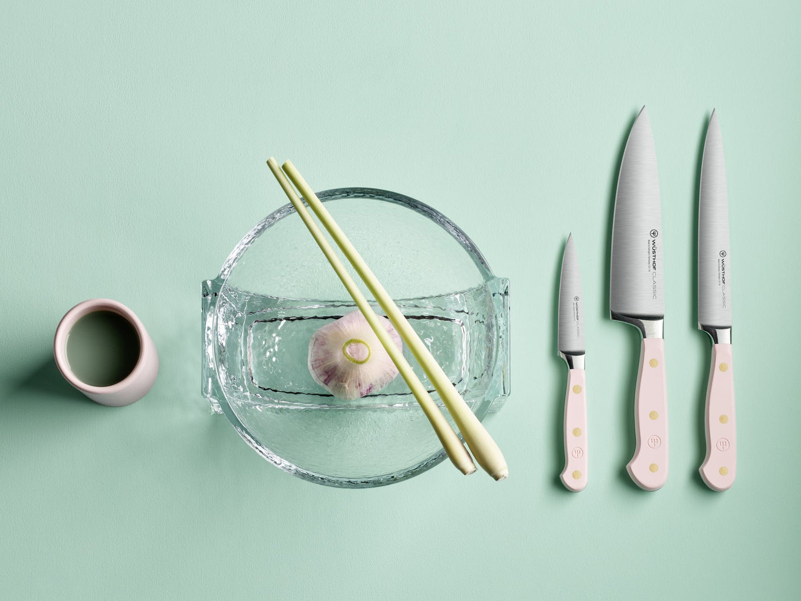 Three Wusthof Classic knives with pink handles and silver blades alongside a glass bowl with garlic and lemongrass