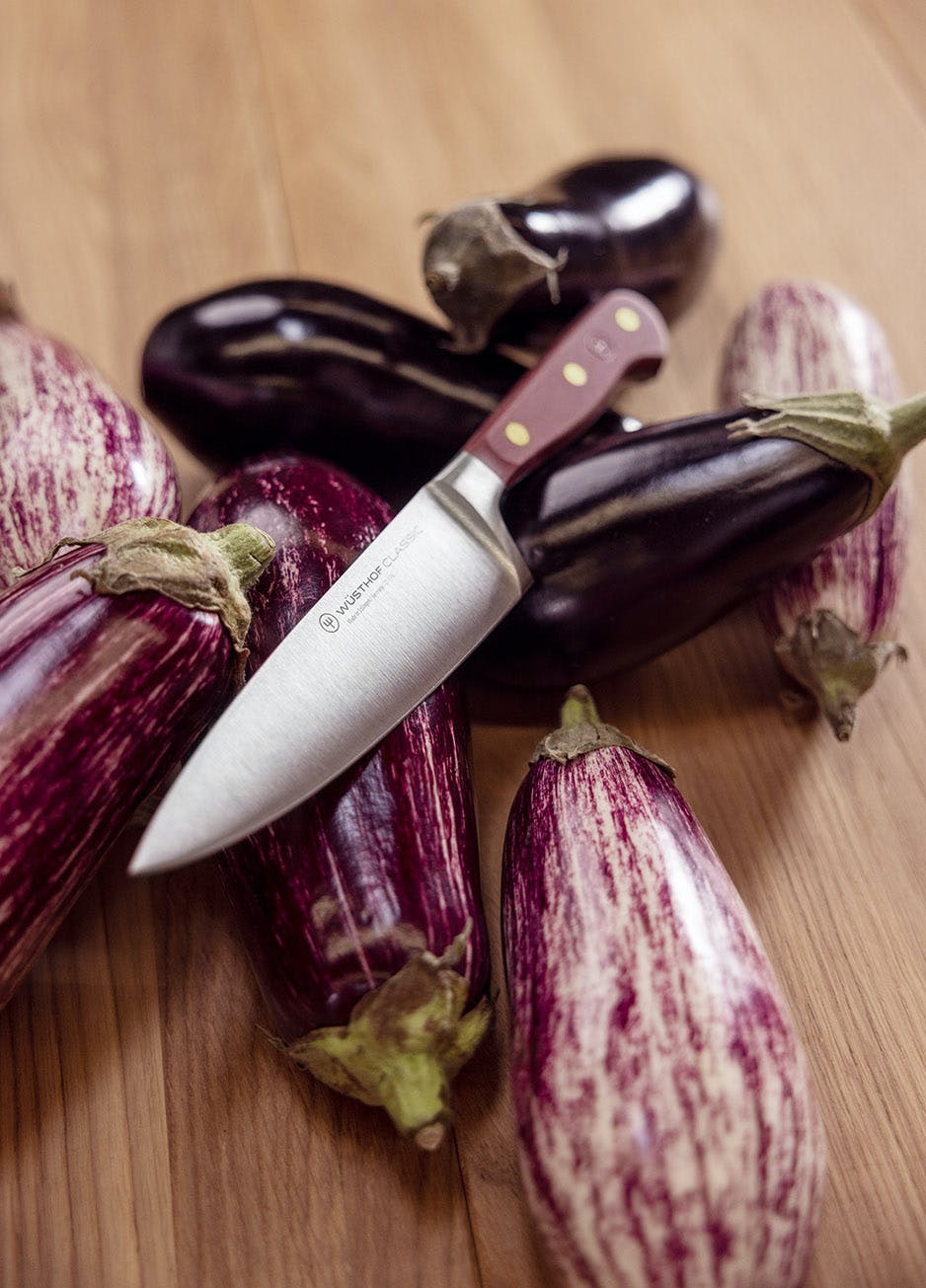 Wusthof Classic 8 Chefs Knife with a sumac-colored handle lying on a light wood surface surrounded by eggplants