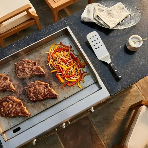 Stainless steel outdoor griddle with four steaks and colorful bell peppers cooking next to a metal spatula