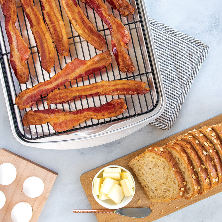 Crispy bacon slices on a roasting rack in a pan next to sliced bread with butter and eggs on cutting boards