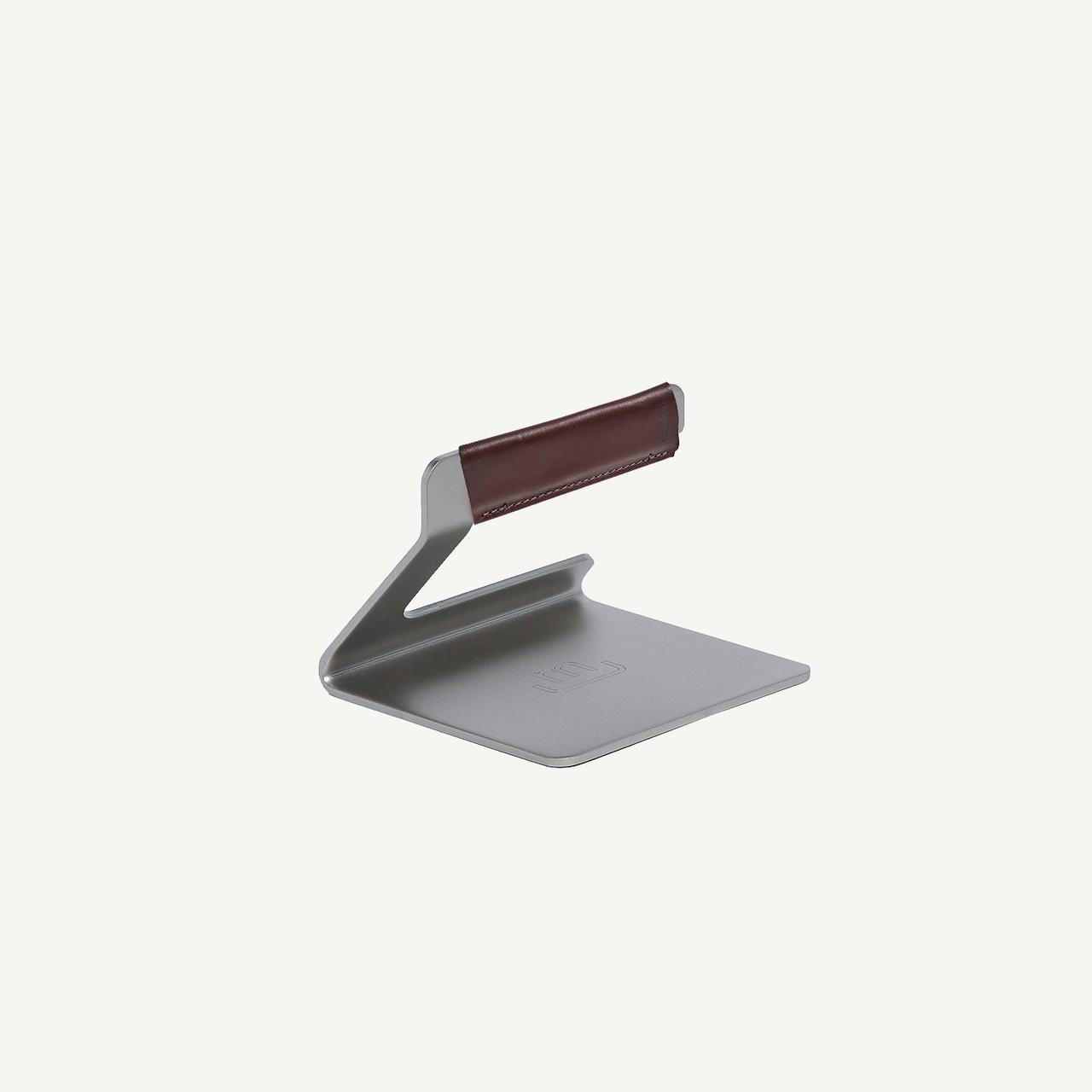 Stainless steel grill press with a flat square base and an angled handle wrapped in dark brown leather