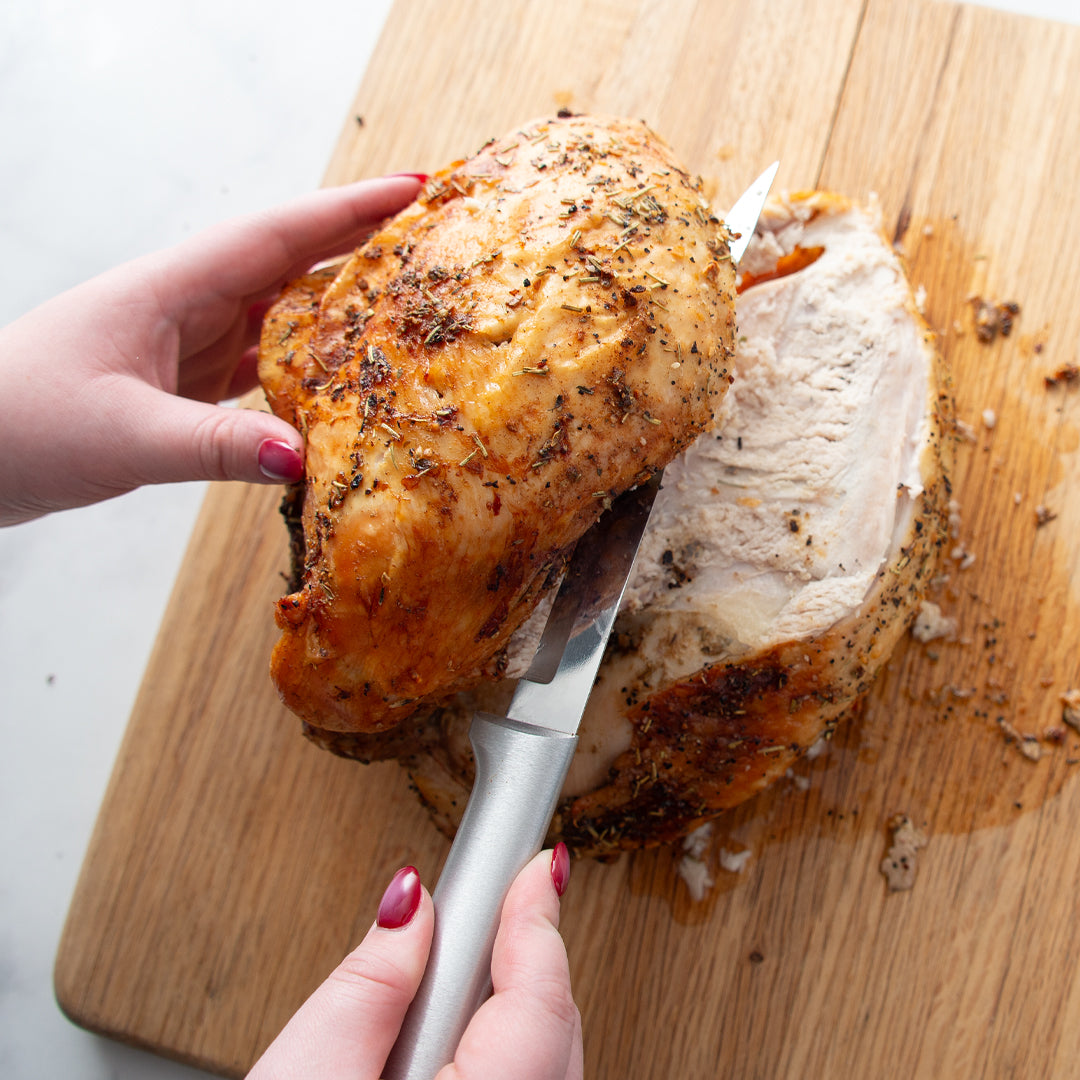 Hands carving a golden-brown herb-crusted turkey breast on a light wooden cutting board with a silver knife