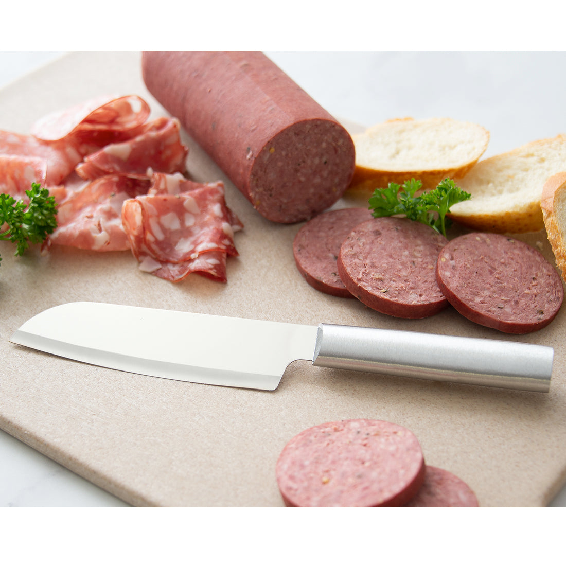 Stainless steel santoku knife on a cutting board surrounded by sliced sausage and bread