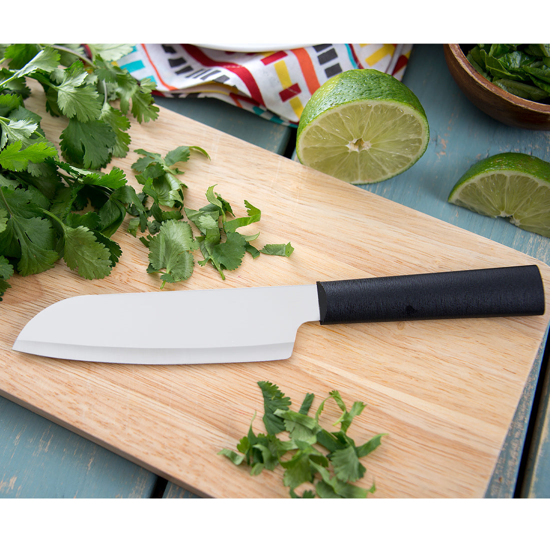White ceramic santoku knife with a black handle on a wooden cutting board surrounded by cilantro and limes