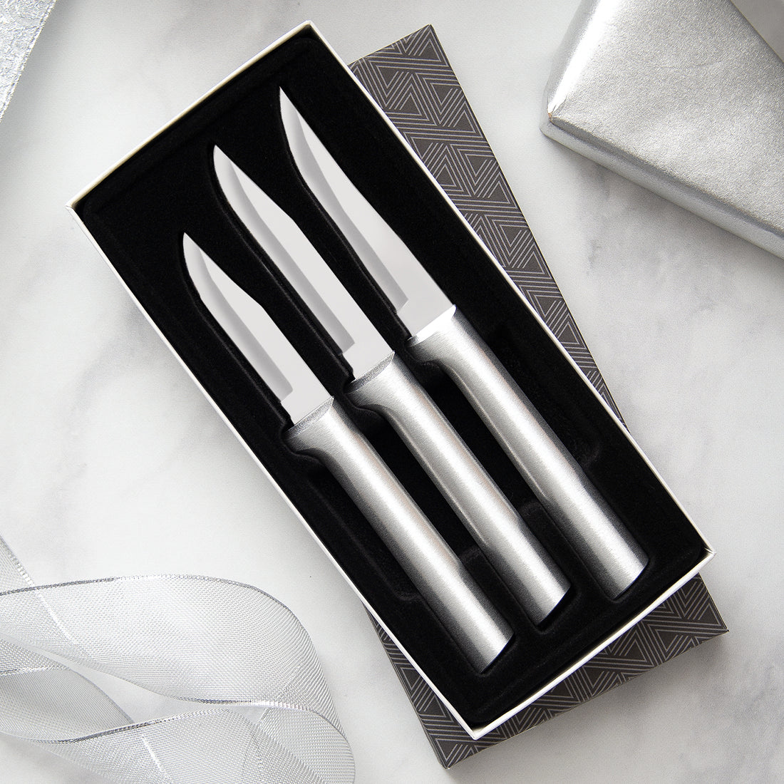 Three stainless steel steak knives in a black and white geometric pattern gift box angled with ribbon