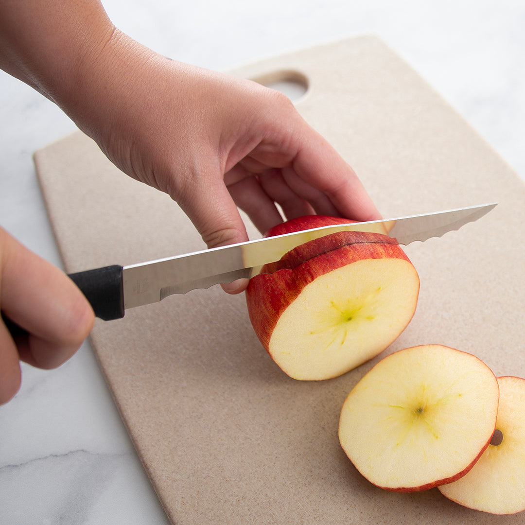 Serrated stainless steel knife with black handle slicing a red apple on a tan cutting board