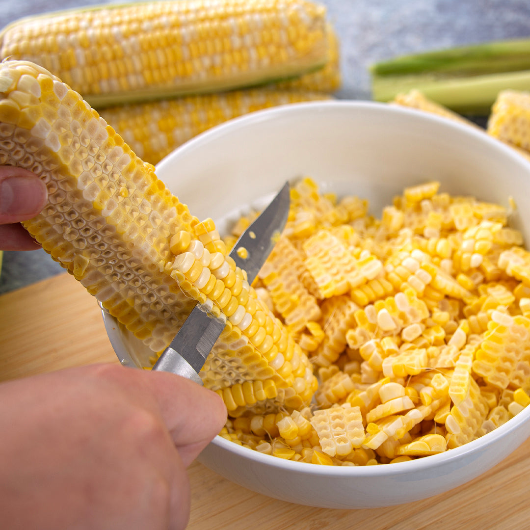 Hand using a specialized tool to remove kernels from a yellow corn cob into a white bowl filled with kernels