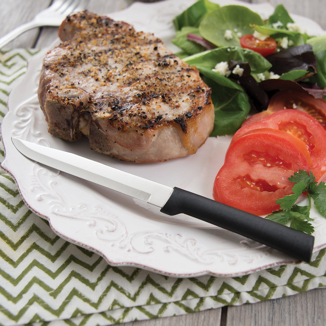 Silver steel steak knife with a black plastic handle resting on a white plate with steak and vegetables