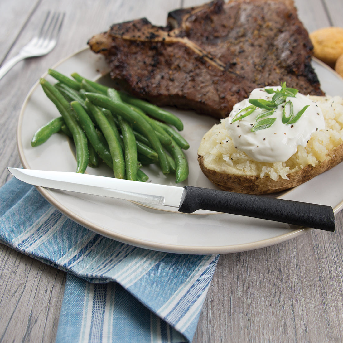 Silver steel steak knife with a black plastic handle resting on a white plate with steak and vegetables