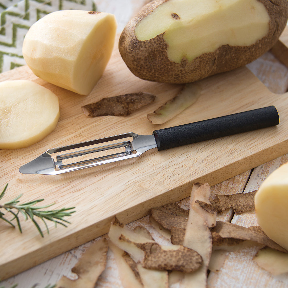 Stainless steel vegetable peeler with black handle used to peel potatoes on a wooden cutting board